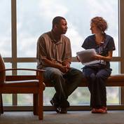 A nurse in blue scrubs holding papers in her hand talks with a male patient in a clinic waiting room