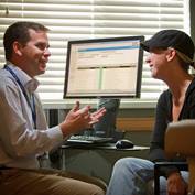 A male doctor sitting in front of a computer talks with a female patient wearing a black baseball cap