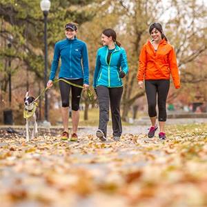 Three young women and a dog are walking together down a leafy sidewalk