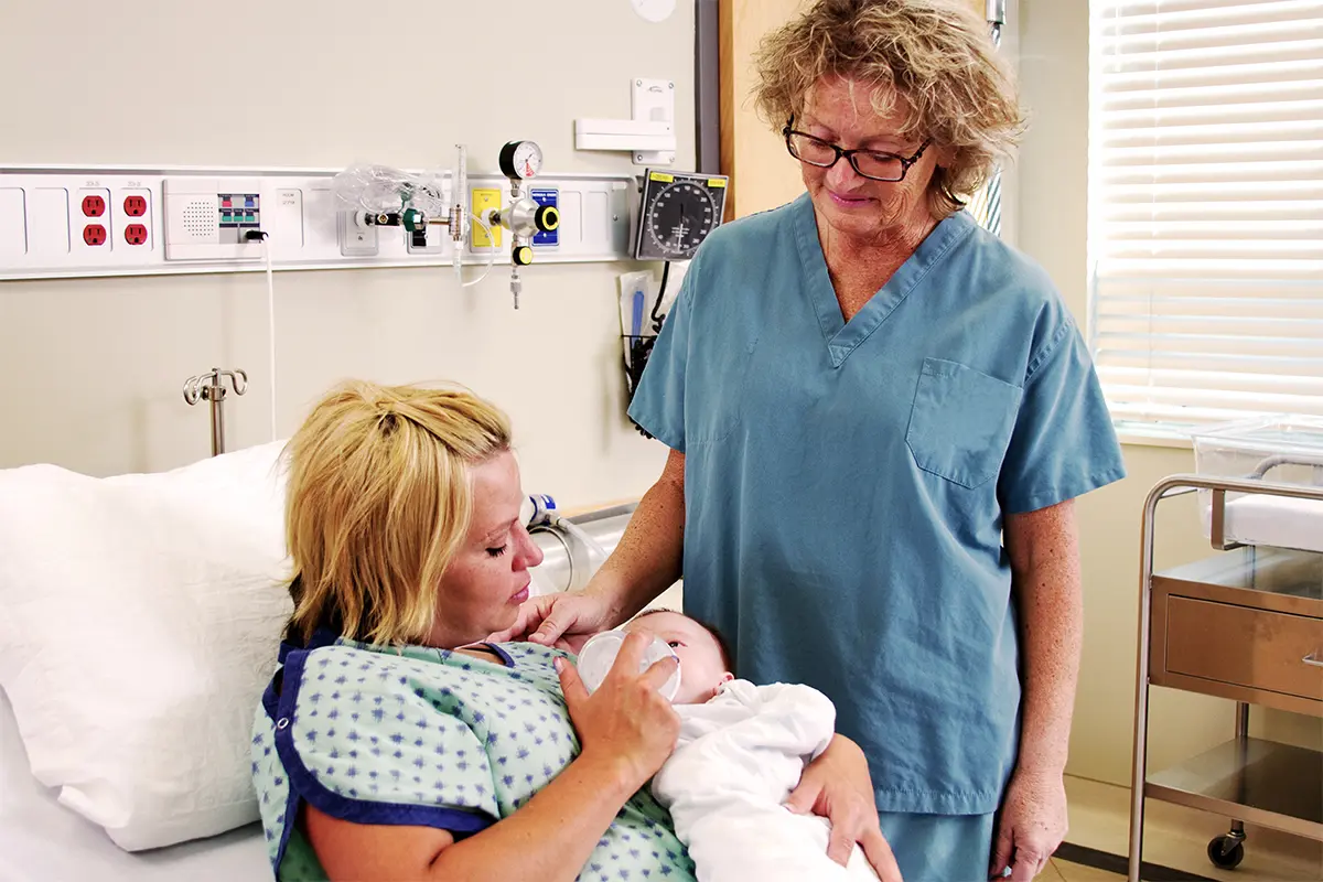 Mother in a hospital bed cradling a newborn with a medical professional next to the bed