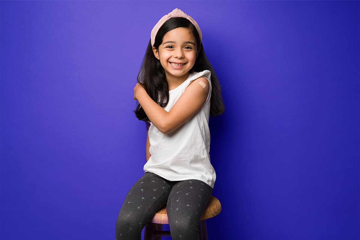 Young girl with a bandaid on her shoulder sitting on a stool against a purple background