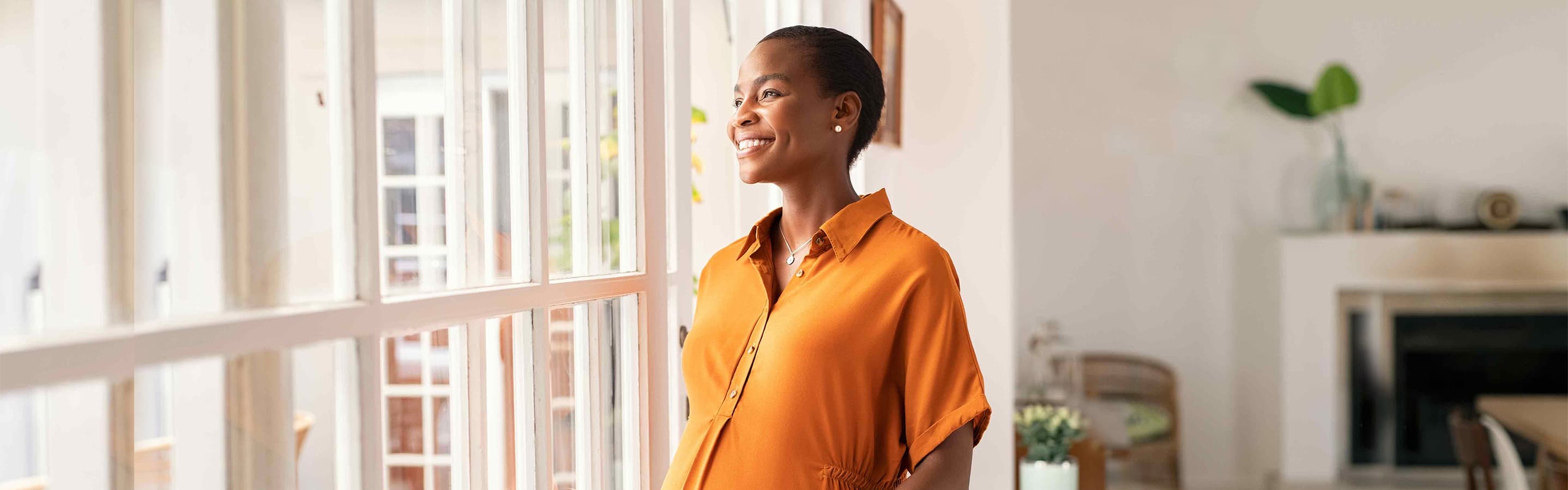 Pregnant woman in an orange dress looking out of the window smiling