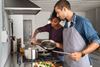 happy younger adult multicultural couple cooking in kitchen over the stove adding ingredients to a pot